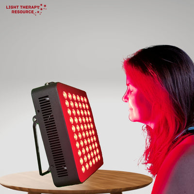 A lady sitting at a table getting a red light therapy session on her face from a red and infrared light therapy tabletop panel