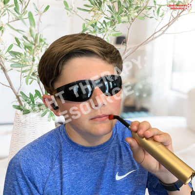 A boy wearing safety glasses and holding a red light therapy handheld torch against his face near his lip for a sore with plants in the background